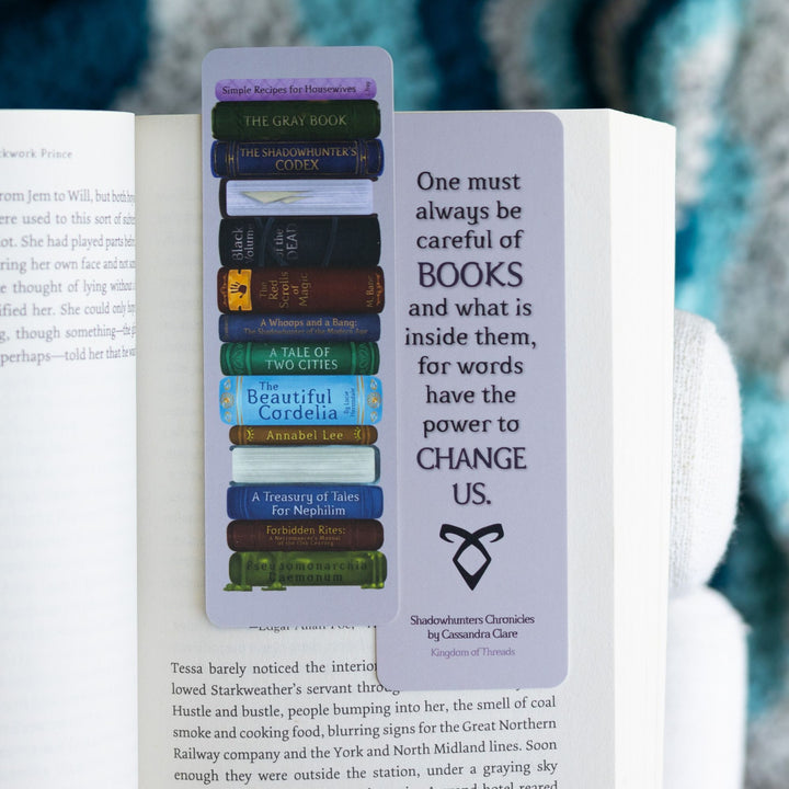 Two purple bookmarks in an open book. One side is stack of books from the Shadowhunters world. Other side reads ‘One must always be careful of books and what is inside them, for words have the power to change us.’
