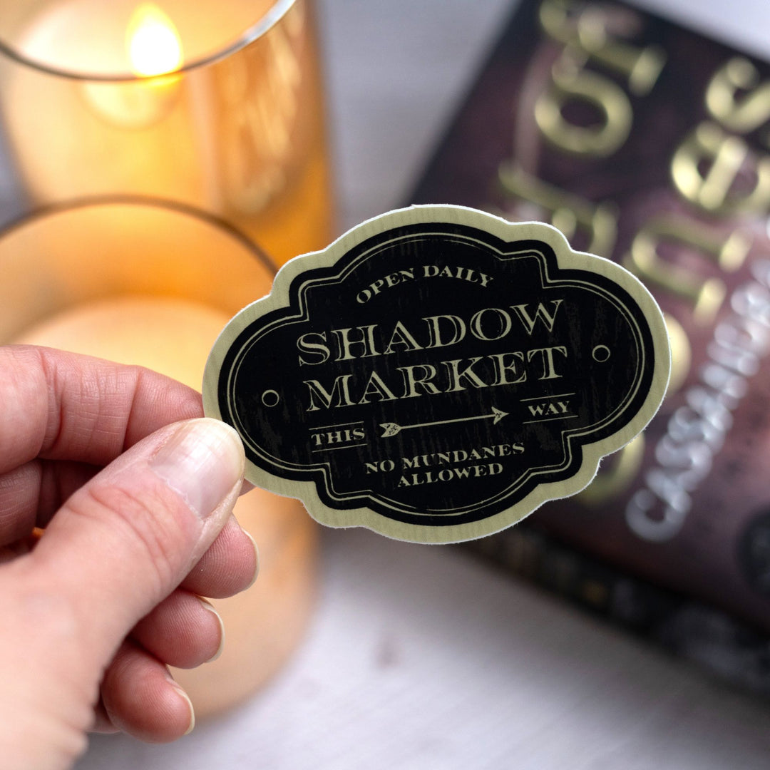 Sticker looks like an antique sign and reads ‘Open daily. Shadow Market, this way. No mundanes allowed’ held over City of Bones.
