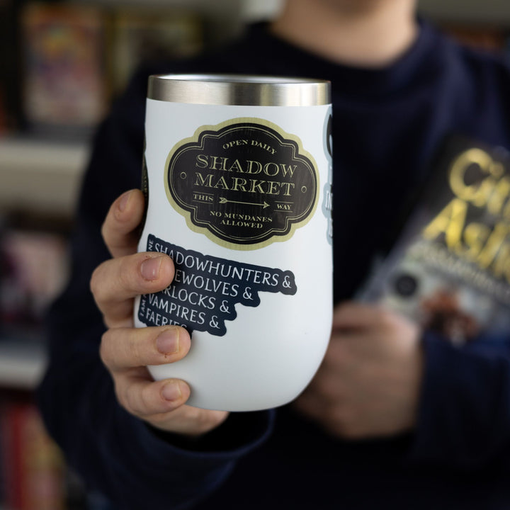 Sticker on a white tumbler, looks like an antique sign and reads ‘Open daily. Shadow Market, this way. No mundanes allowed.’