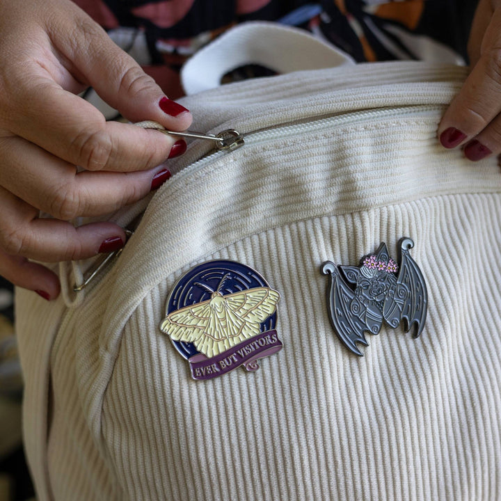 Circular enamel pin inspired by The Knight and the Moth pinned to a corduroy backpack alongside a pin of a grey gargoyle. The pin is a moth flying on a blue sky over a ribbon banner with the quote ‘Ever But Visitors.’