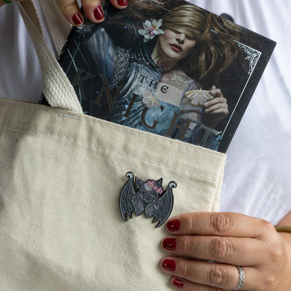 Lifestyle shot of enamel pin of a gargoyle in a flower crown inspired by The Knight and the Moth pinned to a white canvas bag with a book being pulled out of it. The gargoyle is cradling his chin in his paws and smiling.