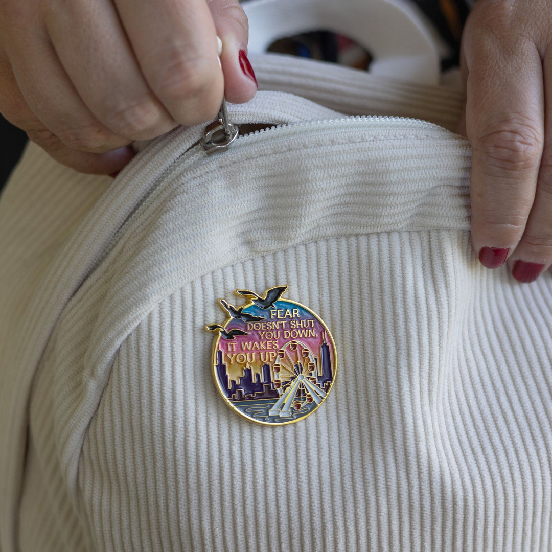 Lifestyle photo of Divergent inspired enamel pin of the Navy Pier Ferris wheel at sunset with the Divergent quote ‘Fear doesn’t shut you down, it wakes you up.’ in gold metal. Pin is on a cream corduroy backpack. Someone is zipping the backpack.