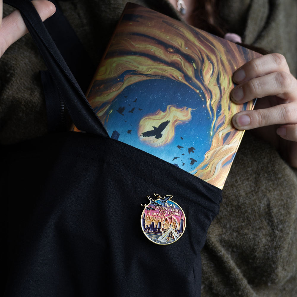 Lifestyle photo of Divergent inspired enamel pin of the Navy Pier Ferris wheel at sunset with the Divergent quote ‘Fear doesn’t shut you down, it wakes you up.’ in gold metal. Pin is on a black bag, held by someone.