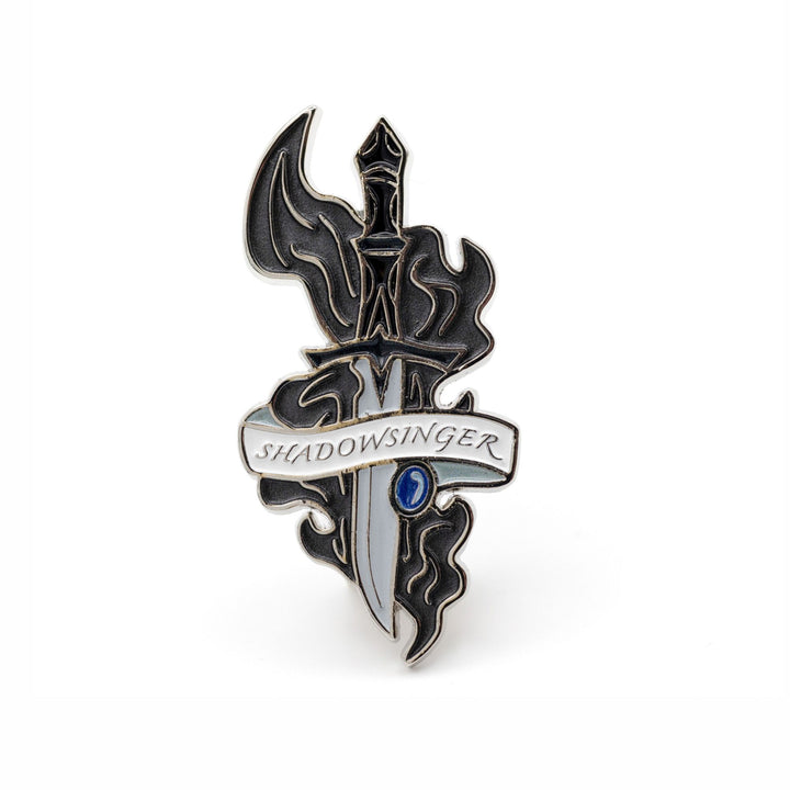 Enamel pin of a dagger with a sapphire stone by the blade and a white ribbon wrapped around it that reads ‘Shadowsinger’ from ACOTAR™. 