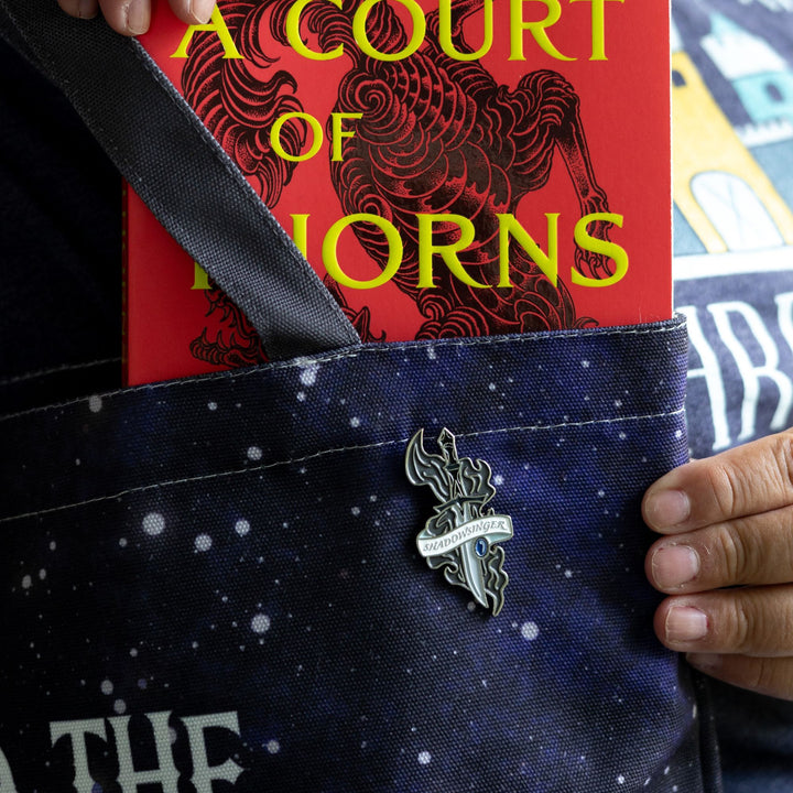 Enamel pin of a dagger with a sapphire stone by the blade and a white ribbon wrapped around it that reads ‘Shadowsinger’ from ACOTAR™, on a white backpack with other ACOTAR™ pins.
