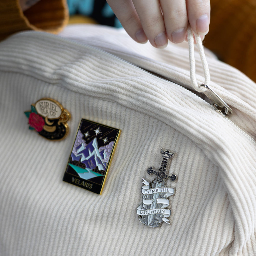 Enamel pin of a sword with a sliced white ribbon around it that reads ‘ Climb the mountain’, on a white backpack with other ACOTAR pins.