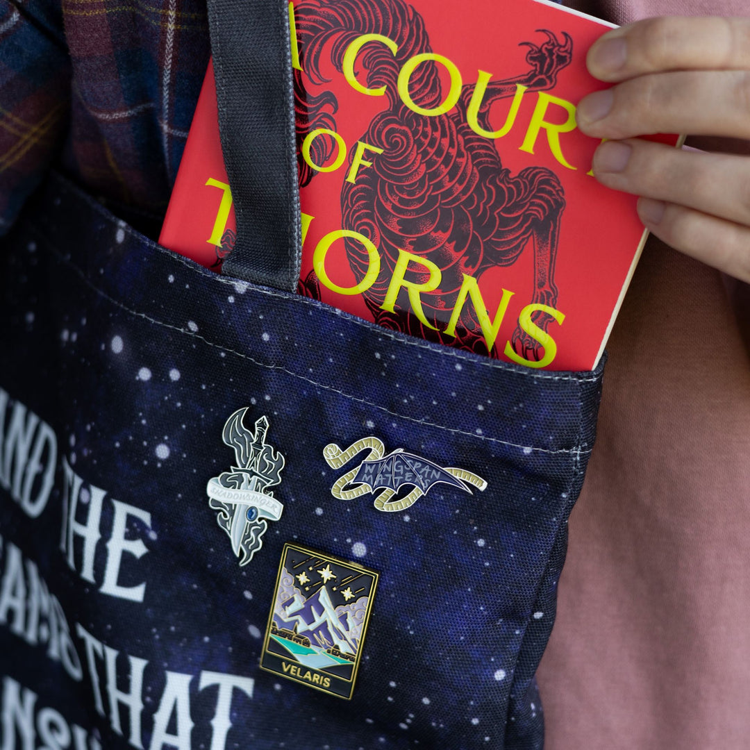 Enamel pin of a bat wing with measuring tape around it and on the wing it reads ‘wingspan matters’, on a blue bag with other ACOTAR pins.