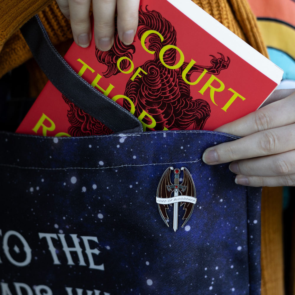 Enamel pin of a sword with a red stone and pommel, surrounded by bat wings that reads ‘Lord of Bloodshed’ on a white ribbon, pinned to a blue bag.