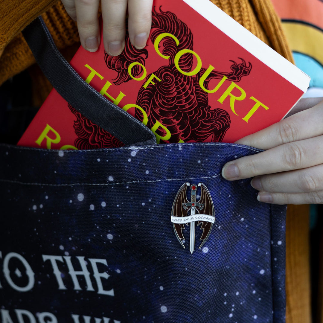Enamel pin of a sword with a red stone and pommel, surrounded by bat wings that reads ‘Lord of Bloodshed’ on a white ribbon, pinned to a blue bag.