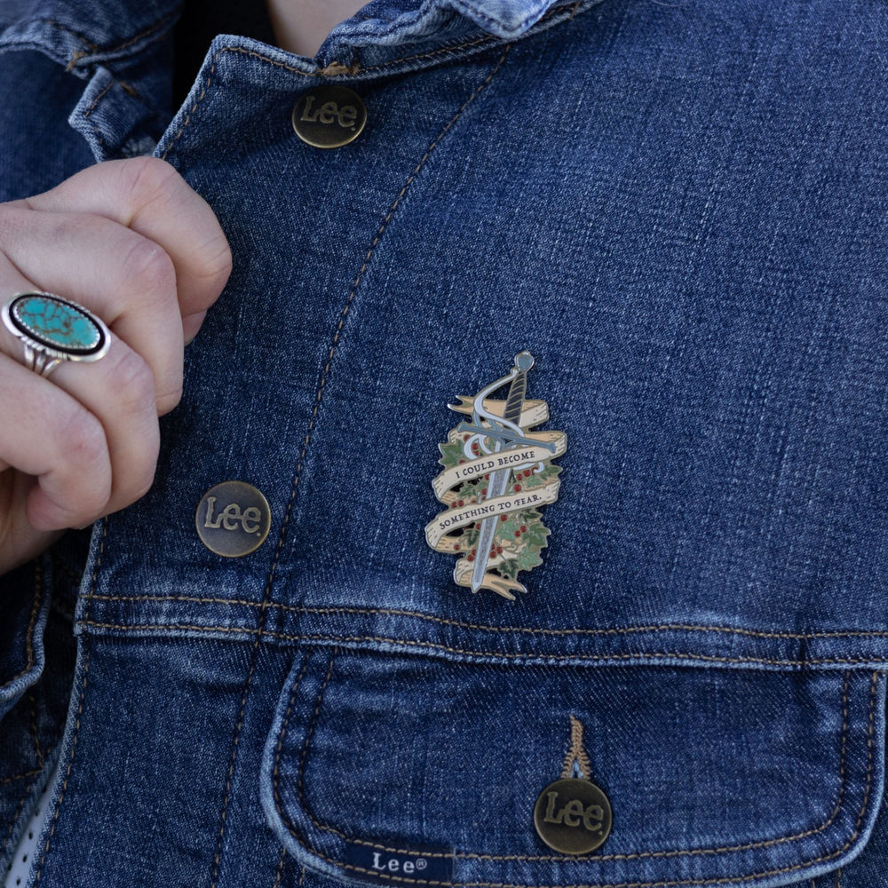 Cruel Prince inspired enamel pin of Jude’s sword Nightfell with banner that reads ‘I could become something to fear’ in gray metal. It is pinned to a jean jacket.
