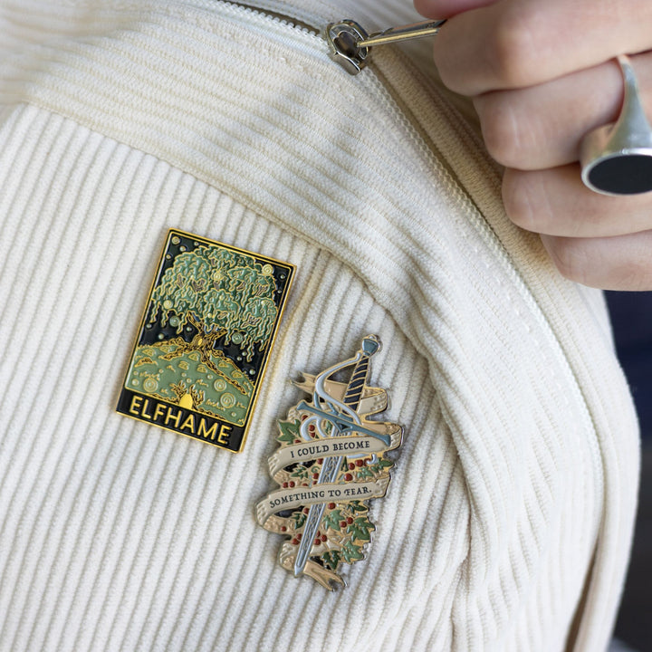 Cruel Prince inspired enamel pin of Jude’s sword Nightfell with banner that reads ‘I could become something to fear’ in gray metal pinned to a corduroy backpack next to another pin that looks like a travel poster and says Elfhame.