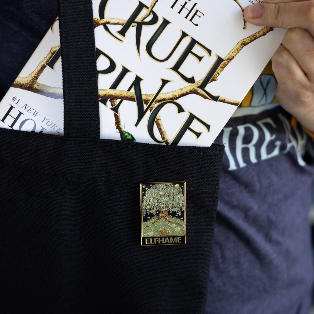 Cruel Prince inspired rectangular enamel pin of Elfhame Palace that reads ‘Elfhame’ in gold metal, pinned to a black tote bag with The Cruel Prince book being put in it.