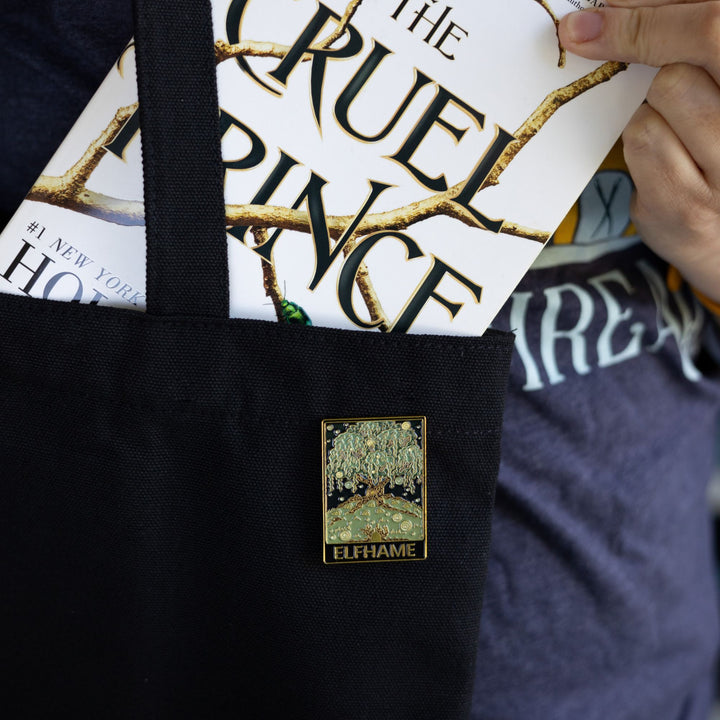 Cruel Prince inspired rectangular enamel pin of Elfhame Palace that reads ‘Elfhame’ in gold metal, pinned to a black tote bag with The Cruel Prince book being put in it.