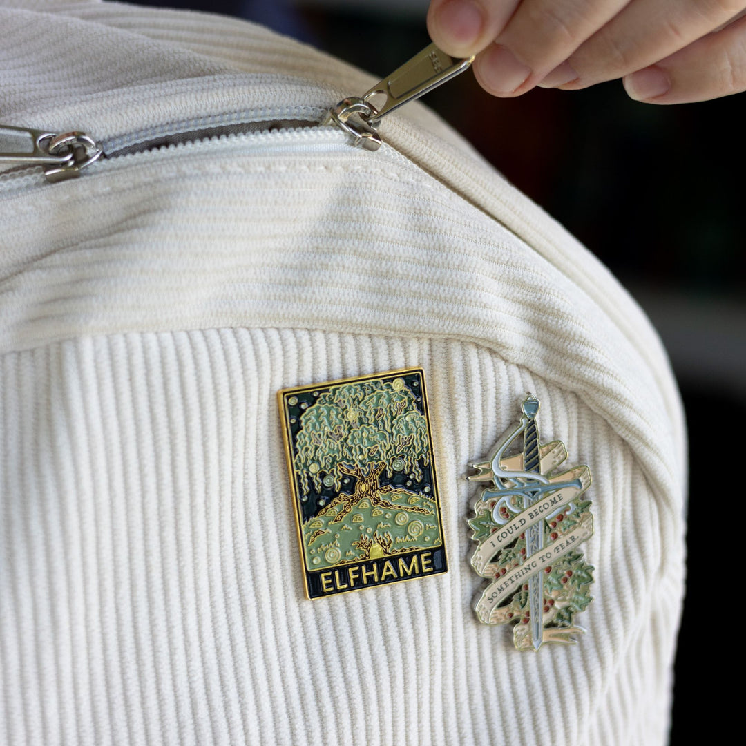 Cruel Prince inspired rectangular enamel pin of Elfhame Palace that reads ‘Elfhame’ in gold metal, pinned to a corduroy backpack next to another pin that is Jude’s sword Nightfell with banner that reads ‘I could become something to fear’ in gray metal. 