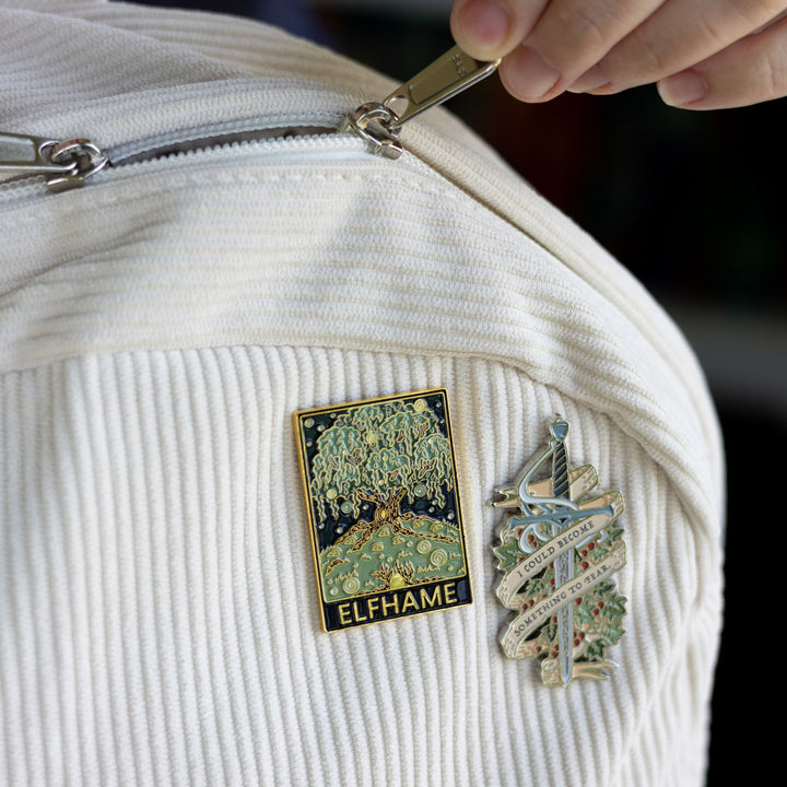 Cruel Prince inspired rectangular enamel pin of Elfhame Palace that reads ‘Elfhame’ in gold metal, pinned to a corduroy backpack next to another pin that is Jude’s sword Nightfell with banner that reads ‘I could become something to fear’ in gray metal. 