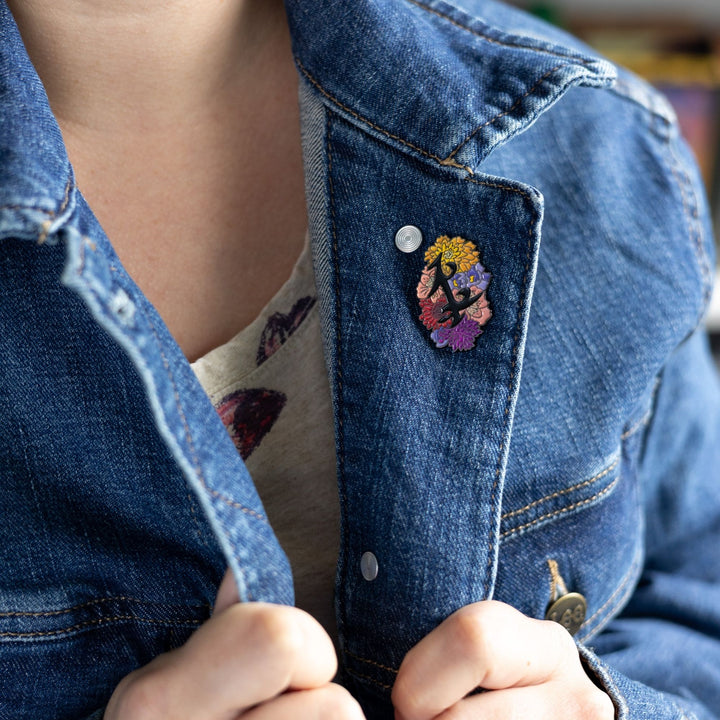 Black enamel pin of parabatai rune from Shadowhunters, surrounded by flowers pinned to a dark blue jean jacket collar.