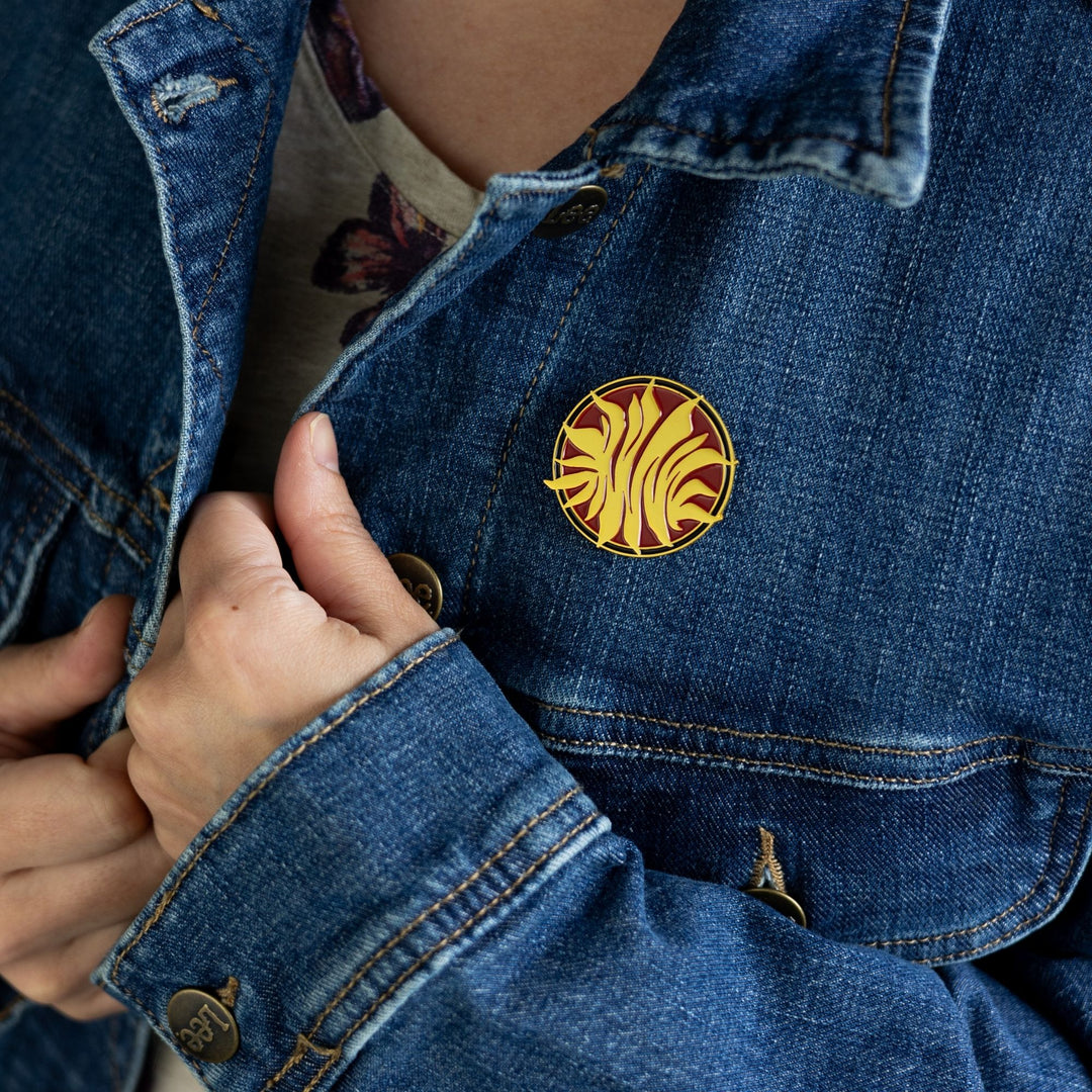 Enamel pin inspired by Red Queen pinned to a jean jacket. The pin is a gold, metal sunburst on a red, circular background.