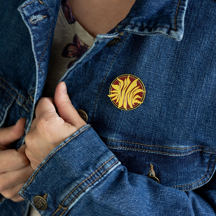 Enamel pin inspired by Red Queen pinned to a jean jacket. The pin is a gold, metal sunburst on a red, circular background.