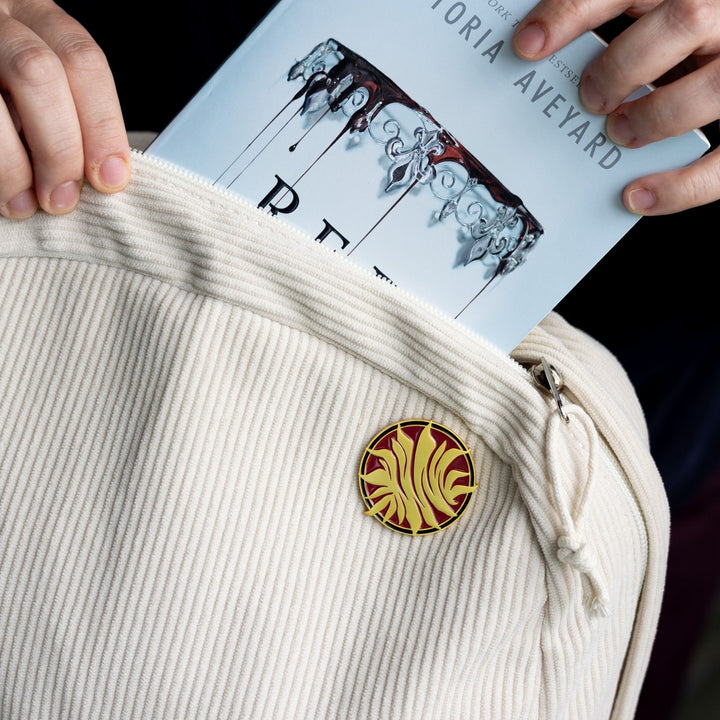 Enamel pin pinned to a corduroy backpack. ARed Queen inspired,  a gold, metal sunburst on a red, circular background. 