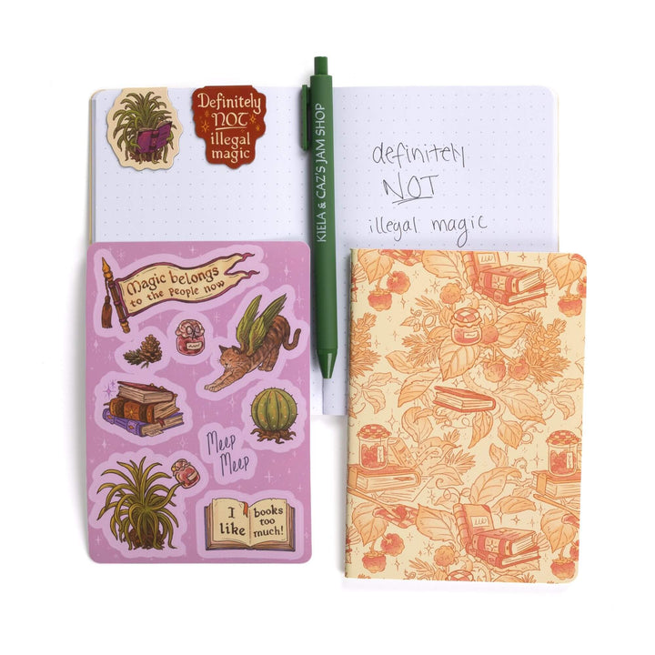 Gift set opened: Dot grid A6 journal open with pen, Caz & “Definitely NOT illegal magic” magnetic bookmarks, Spellshop characters sticker sheet, A6 strawberry & books journal.
