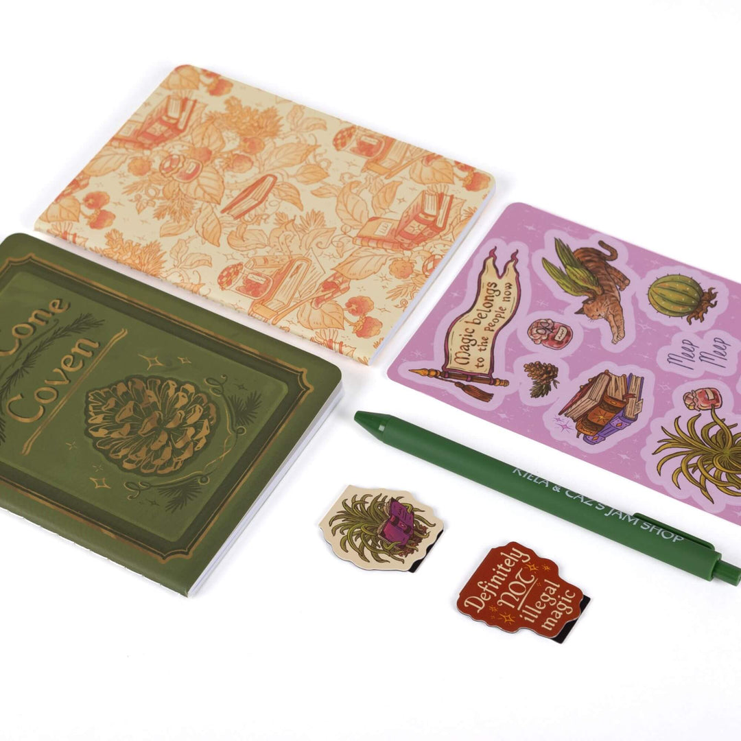 Gift set laid out: A6 “Pine Cone Coven” and strawberry & books journals, Spellshop characters sticker sheet, Caz & “Definitely NOT illegal magic” magnetic bookmarks, green pen.