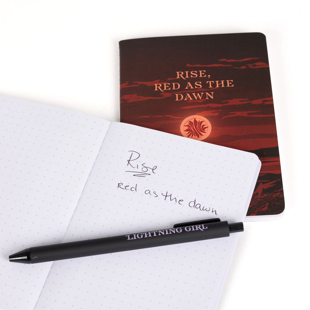 ‘Rise, Red as the Dawn’ A6 journal behind an open A6 dot grid journal with purple pen and handwritten note: ‘Lightning has no mercy.’
