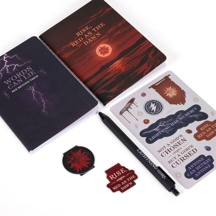 : Gift set laid out: A6 ‘Words can lie’ and ‘Rise, Red as the dawn,’ Red Queen sticker sheet, ‘Rise, Red as the Dawn’ and the Scarlet Guard insignia magnetic bookmarks, black pen.