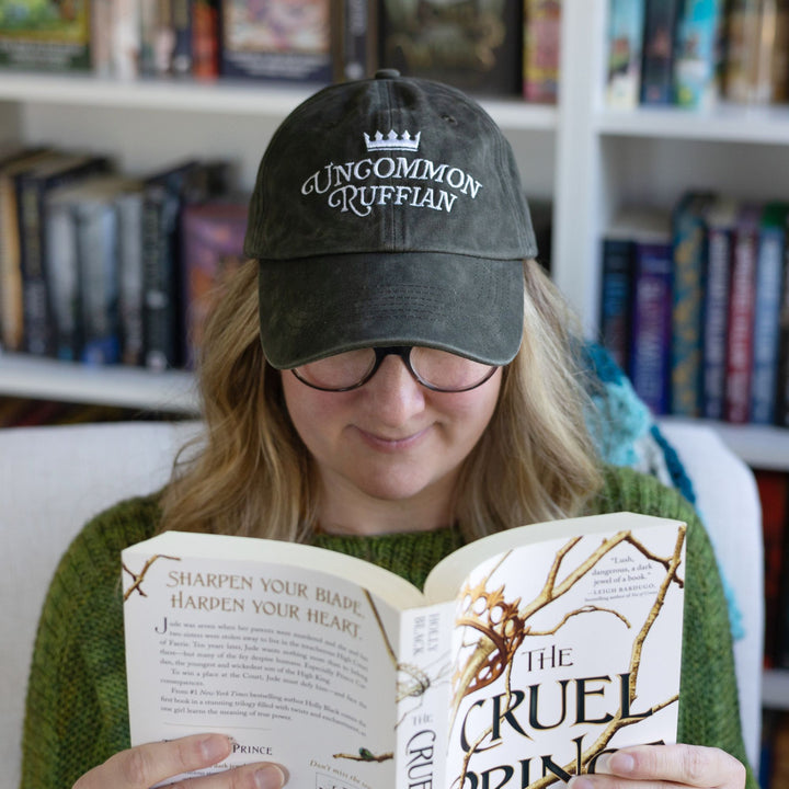 Person wearing dark brownish grey hat with the quote ‘Uncommon Ruffian’  from Holly Black’s Folk of Air series, while reading The Cruel Prince.