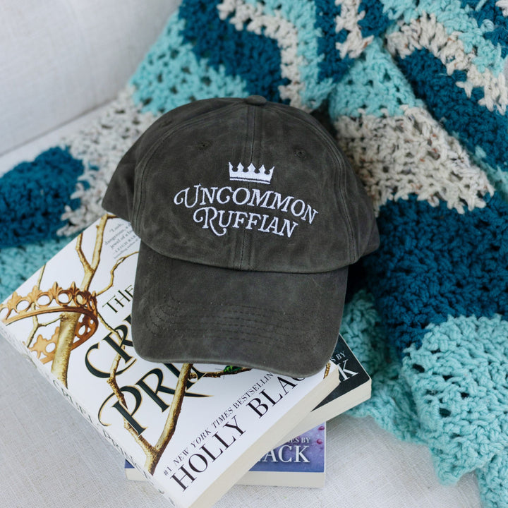 Dark brownish grey hat with the quote ‘Uncommon Ruffian’ on the front from Holly Black’s Folk of Air series on a chair on top of the book series.