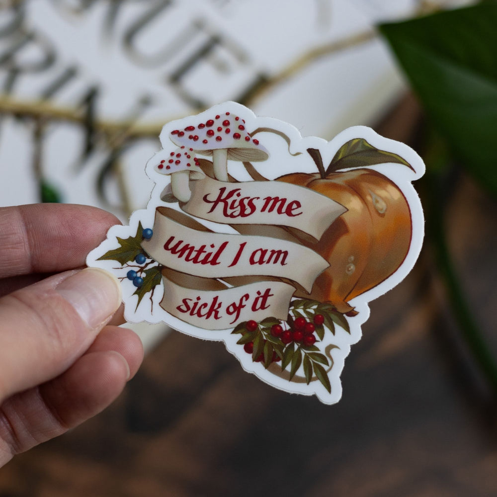 Cruel Prince quote sticker: ‘Kiss me until I am sick of it’ with rowan berries, blusher mushrooms, and faerie fruit. Held above The Cruel Prince book.