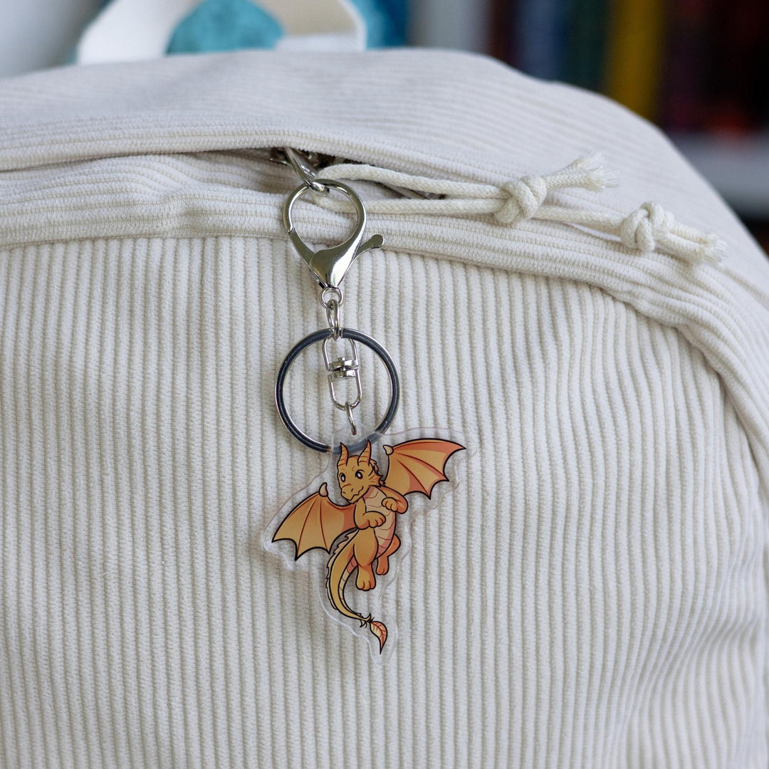 : Acrylic keychain of baby Andarna, a golden dragon, from Fourth Wing by Rebecca Yarros on the clipped to a white backpack.