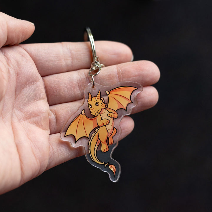 Acrylic keychain of baby Andarna, a golden dragon, from Fourth Wing by Rebecca Yarros held on a palm.
