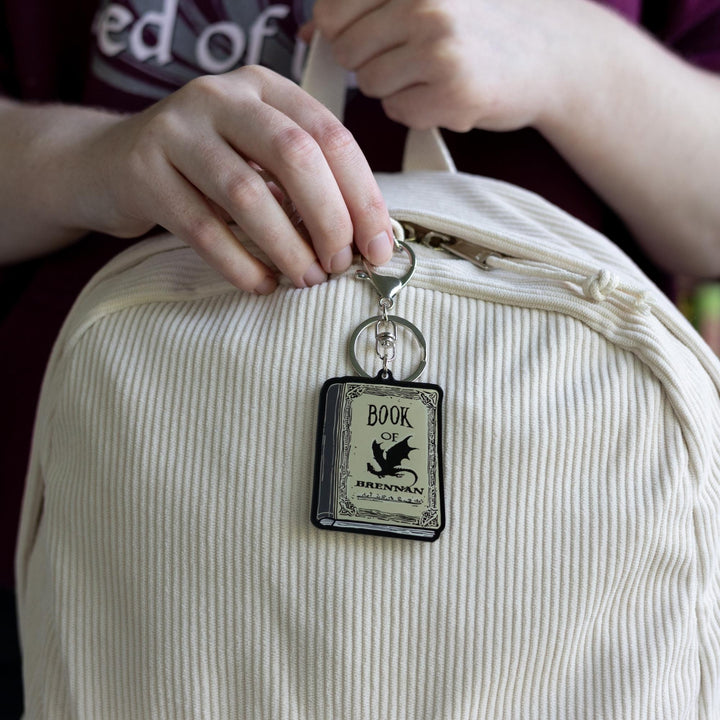  Acrylic keychain of the Book of Brennan from Fourth Wing. The book is parchment color, with black title text and a black dragon on the cover clipped to a white backpack.
