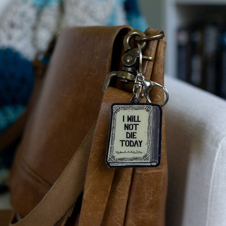  Acrylic keychain of a parchment colored book with black text reading 'I will not die today.' clipped to a leather purse.
