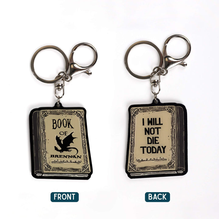 2 acrylic keychains, one of the Book of Brennan and the reverse side, the quote 'I will not die today.' Book is parchment color with black text.