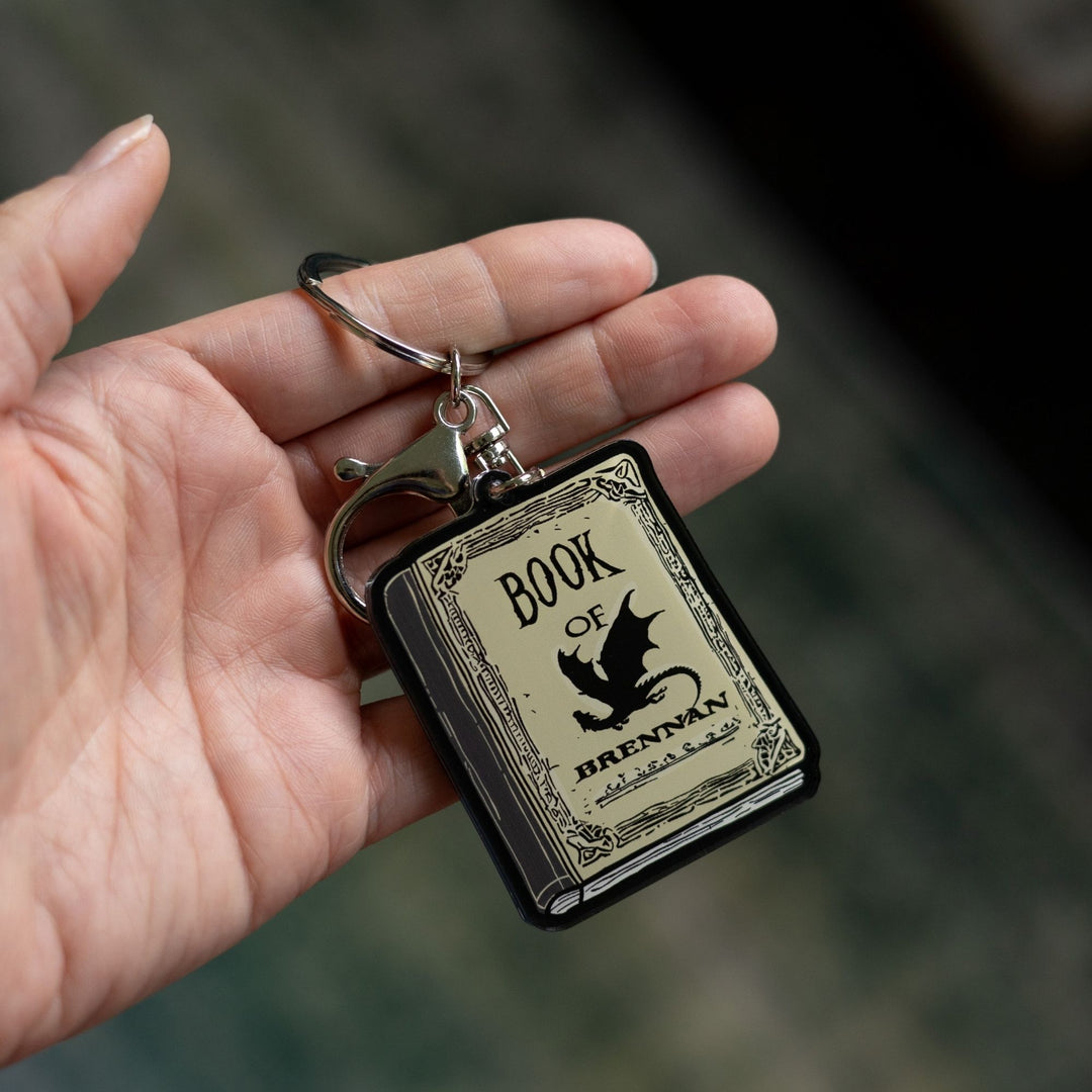 Acrylic keychain of the Book of Brennan, parchment colored, with black title text and a black dragon on the cover held on a palm. 