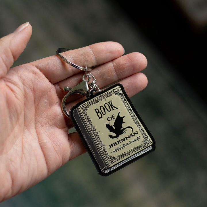 Acrylic keychain of the Book of Brennan, parchment colored, with black title text and a black dragon on the cover held on a palm. 
