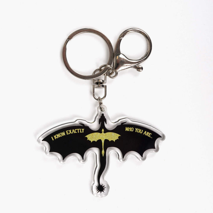 Acrylic keychain of the silhouette of a black dragon, wings spread, with a smaller glittery gold dragon overlaid. Quote on the wings is 'I know exactly who you are.'