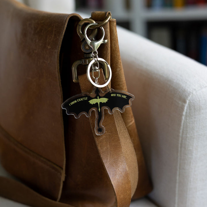 crylic keychain of the silhouette of a black dragon, wings spread, with a smaller glittery gold dragon overlaid clipped to a leather purse. Quote on the wings is 'I know exactly who you are.'
