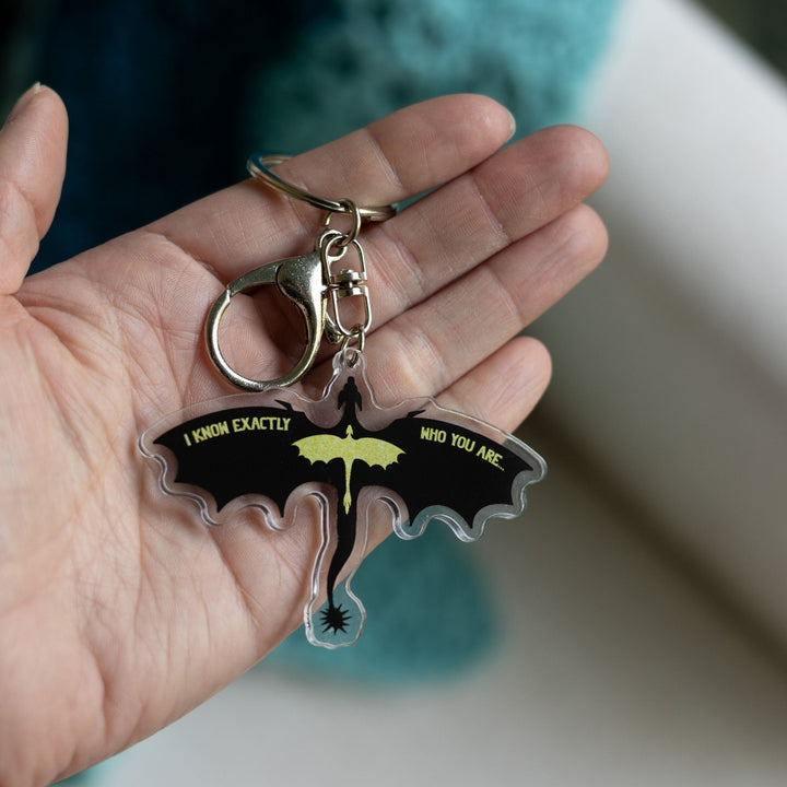 Acrylic keychain of the silhouette of a black dragon, wings spread, with a smaller glittery gold dragon overlaid, held on a palm. Quote on the wings is 'I know exactly who you are.’ 