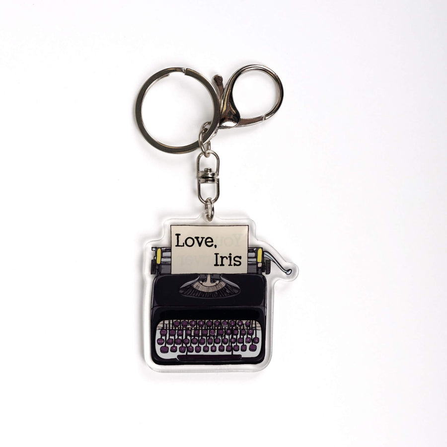  Acrylic keychain of a black typewriter with purple keys. It has a piece of paper in it that says 'Love, Iris.' From Divine Rivals by Rebecca Ross.