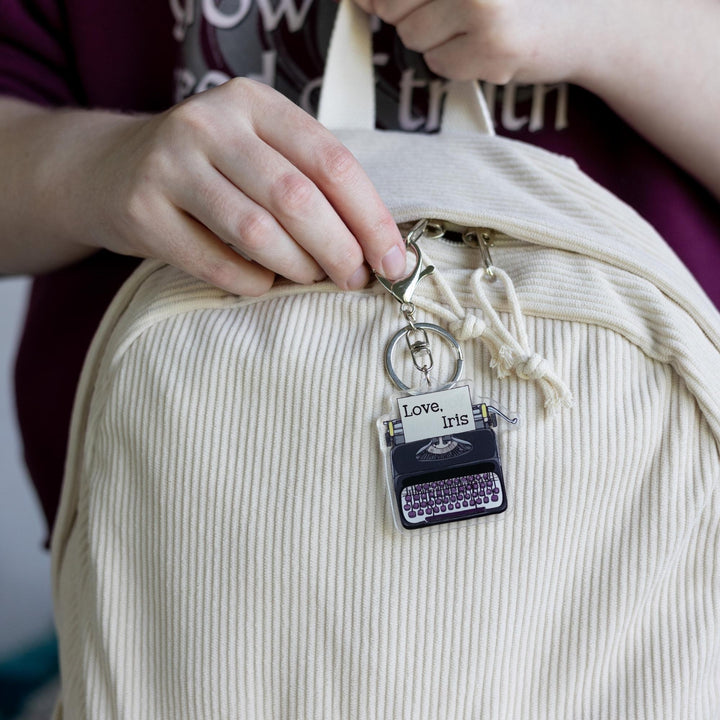  Acrylic keychain of a black typewriter with purple keys clipped to a white backpack. It has a piece of paper in it that says, 'Love, Iris.' From Divine Rivals. 
