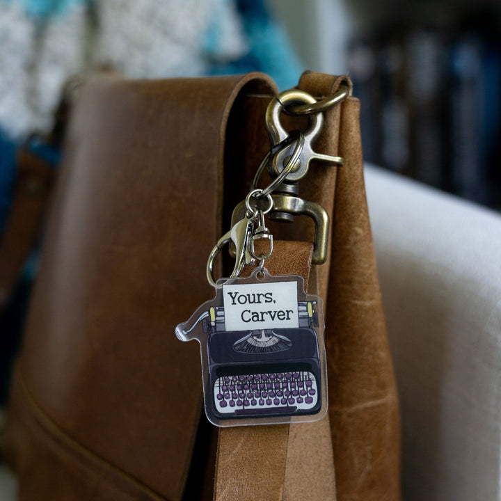 Acrylic keychain of a black typewriter with purple keys clipped to a leather purse. It has a piece of paper in it that says, 'Love, Carver.'  
