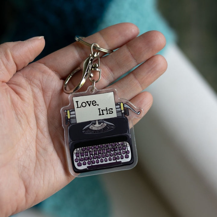 Acrylic keychain of a black typewriter with purple keys held on a palm. It has a piece of paper in it that says 'Love, Iris.’