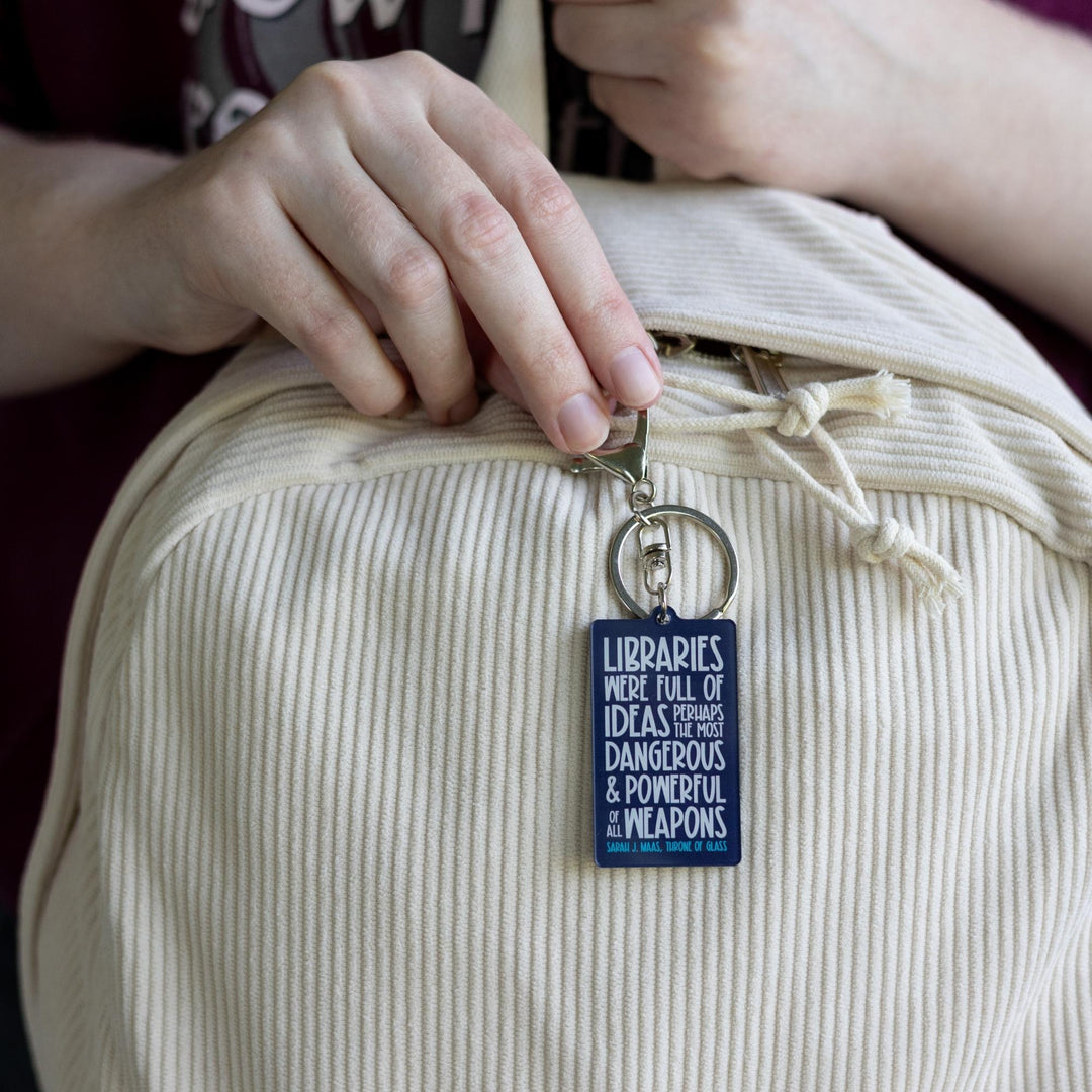  Dark blue acrylic keychain 'Libraries were full of ideas, perhaps the most dangerous and powerful weapons of them all. Sarah J. Maas Throne of Glass’  clipped to a white backpack.
