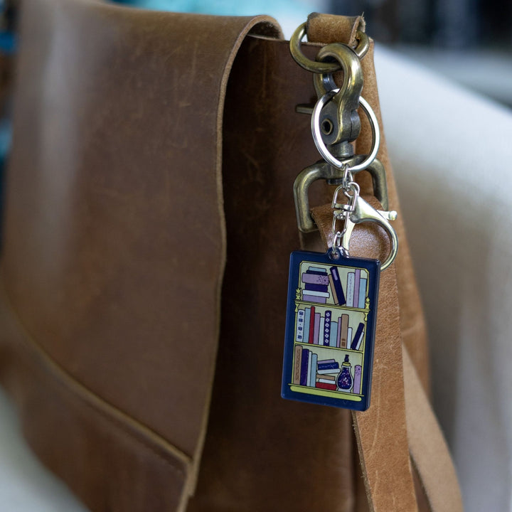 Dark blue acrylic keychain of a full bookshelf with fun spines clipped to a leather purse.