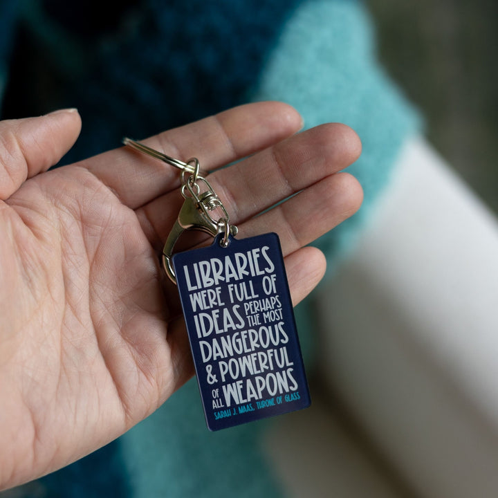 Dark blue acrylic keychain. 'Libraries were full of ideas, perhaps the most dangerous and powerful weapons of them all. Sarah J. Maas Throne of Glass.’ Held on a palm.
