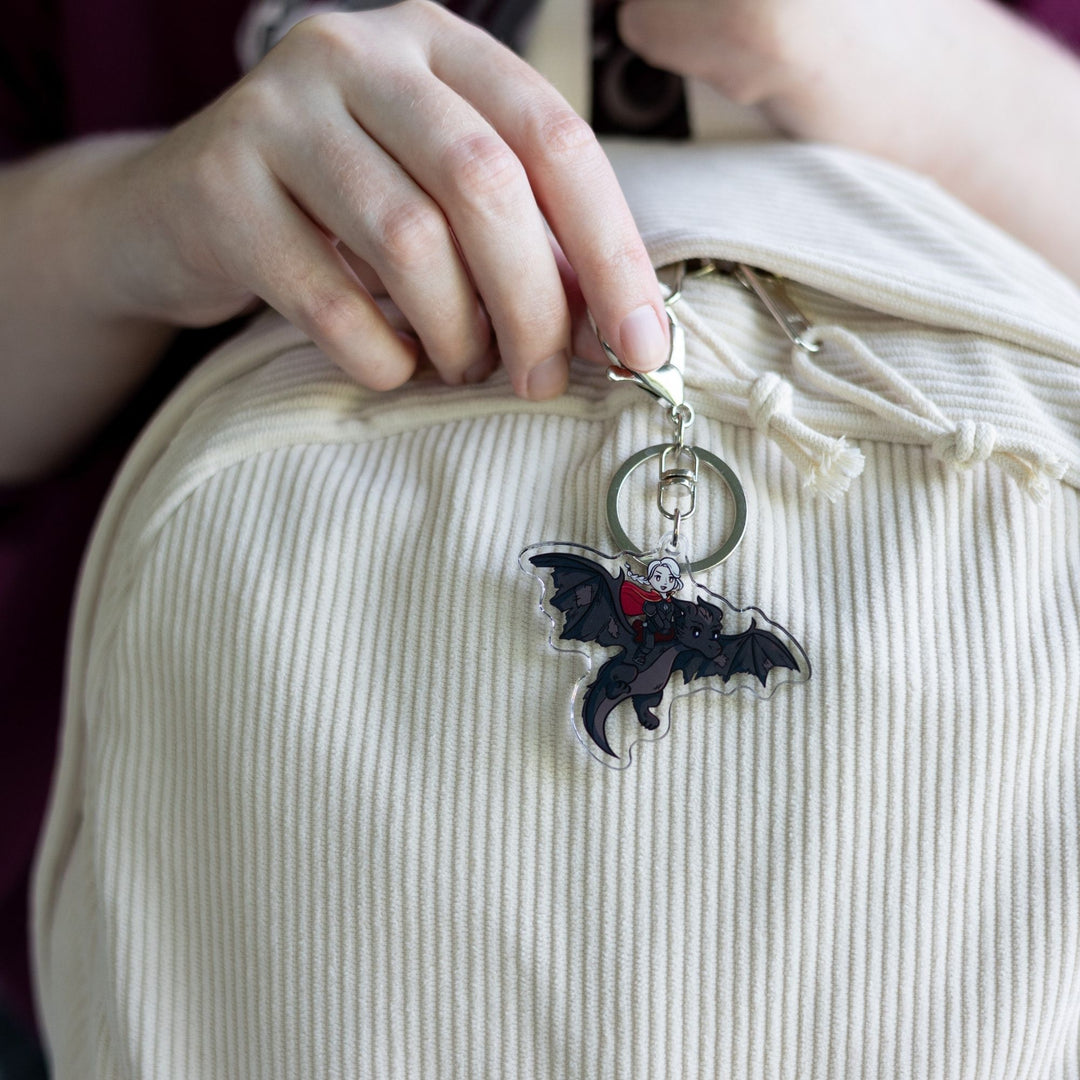  Acrylic keychain of a cartoon of the witch Manon riding Abraxos, her black wyvern  clipped to a white backpack.