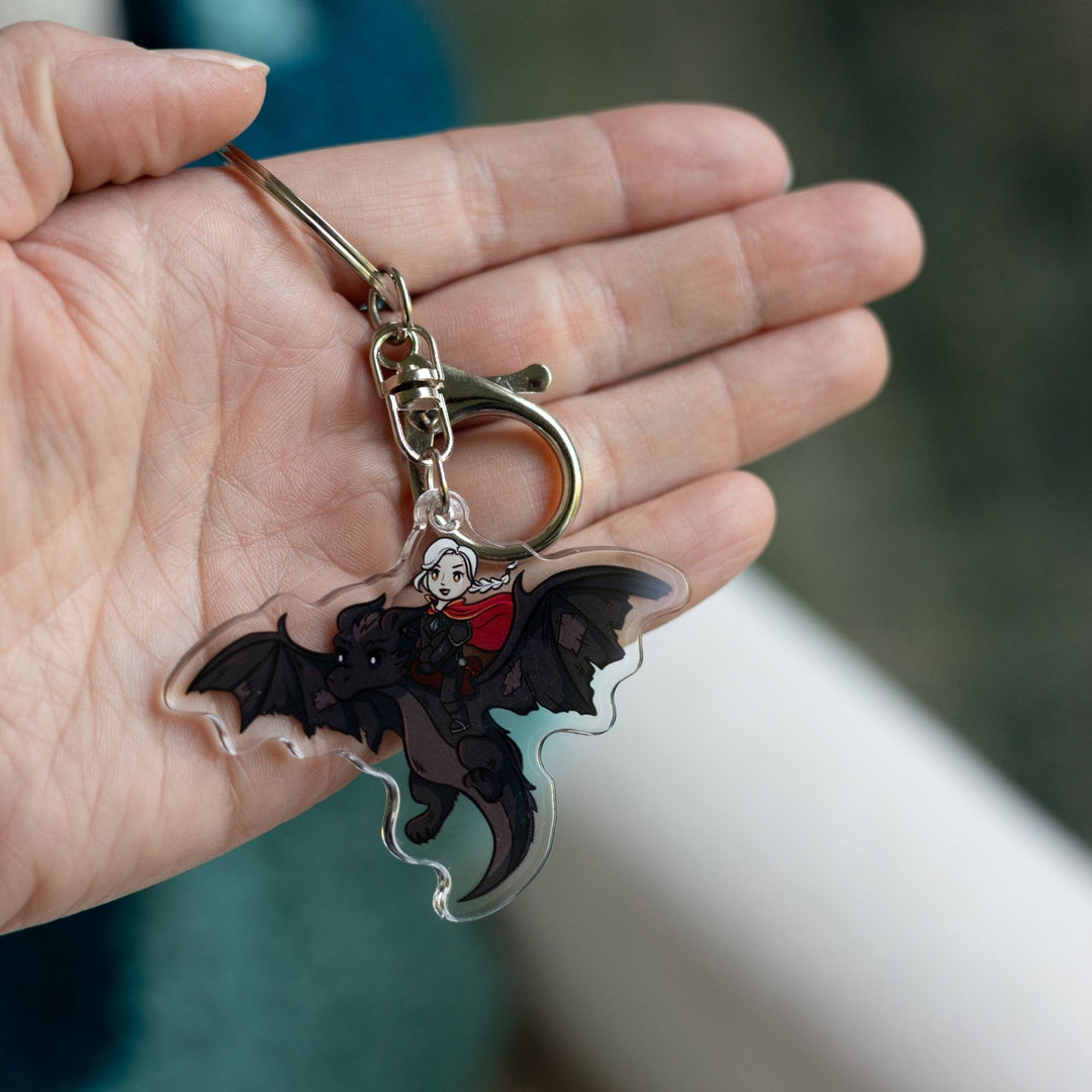 Acrylic keychain of a cartoon of the witch Manon riding Abraxos, her black wyvern, held in a palm.
