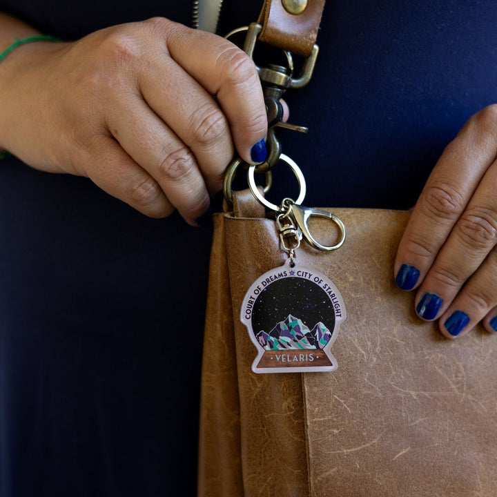 Acrylic keychain of snow globe with mountains against the night skyclipped to a leather purse. 'Court of Dreams City of Starlight' above it. ‘Velaris’ on the base.
